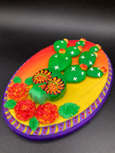 Load image into Gallery viewer, This handcrafted polymer clay wall art features a wood frame painted in a gradient from light yellow to dark red, creating a stunning sunset look. With a bright green skull cactus plant and bright yellow/red blooming flowers. Boasting vivid marigold blooms and a deep purple frame to make a bold contrast with the warm background hue. this piece is sure to make a statement.  Measurements: H 7&quot; x W 5” x D 1 1/2&quot;
