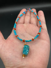 Load image into Gallery viewer, This adjustable choker style necklace features an eye-catching teal wood scarab beetle pendant and glass beaded strands. This dramatic necklace is kept together by double-hole flat matte lava-red beads, providing a collar shape. The necklace artfully wraps around the neck, conveying an elegant and bold style.  Measurements:    Necklace is 14 1/2” in length, can be adjusted up to 16&quot; 
