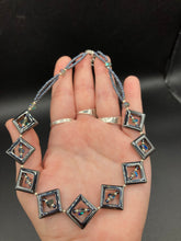 Load image into Gallery viewer, This two-strand necklace, crafted from aqua-blue and silver toned glass beads, is a stunning statement piece. The addition of faceted triangle-shaped glass beads tucked between hematite square beads adds an exquisite and unique flair. The necklace gracefully drapes around the neck, bringing an air of bold and elegant flair to any ensemble it complements. 15” in length, can be extended up to 17"
