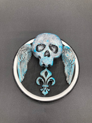 This handcrafted Dia De Los Muertos Calavera wall art decor is a unique way to bring life to your home and celebrates Mexican culture. Made with polymere clay and painted on a wooden frame, the beautiful blue skull is finished with wings and fleur de lis embellishments for a sophisticated look. Perfect for any room!  Measurements: D 4