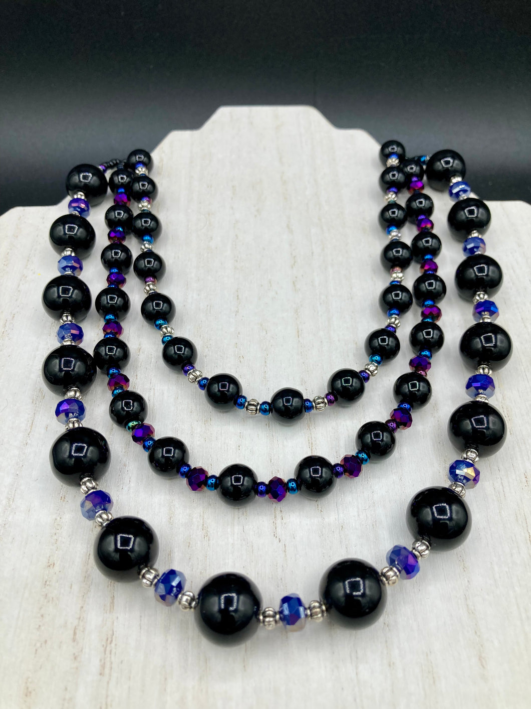 This Bohemian style necklace is composed of three strands of shimmery large black and dark purple glass beads, along with floral metal beads. The elegant drape creates a bold and fierce look that is ideal for a variety of occasions, such as cocktail chic attire and weddings. It is a perfect minimalist accessory.  Measurements:  Necklace is 16” in length - adjustable to 18
