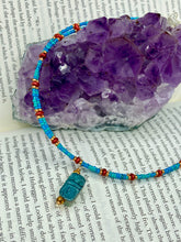 Load image into Gallery viewer, This adjustable choker style necklace features an eye-catching teal wood scarab beetle pendant and glass beaded strands. This dramatic necklace is kept together by double-hole flat matte lava-red beads, providing a collar shape. The necklace artfully wraps around the neck, conveying an elegant and bold style.  Measurements:    Necklace is 14 1/2” in length, can be adjusted up to 16&quot; 
