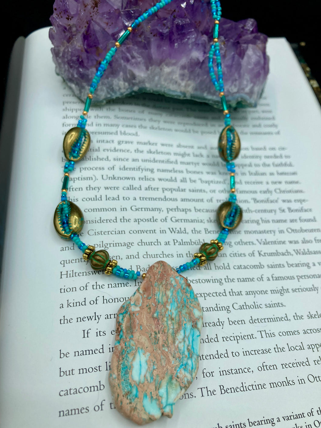 This stunning two-strand necklace features an array of glass beads in hues of green and blue turquoise, gold and green for a vibrant, colorful and bold look. The centerpiece is a large howlite stone chip dyed in turquoise color, accented by the bronze patina cowry beads.  Measurements: Necklace is 17” 