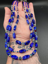 Load image into Gallery viewer, This vibrant royal blue and silver-tone necklace is adjustable and features an eye-catching three-strand design. The rectangular blue glass beads are predominant, complemented by a blend of lighter blue and silver beads for an intriguing multi-hued finish. By mixing shapes and tones, it creates a complex, dimensional look. The draped effect adds a fierce yet elegant pizzazz to any outfit. Perfect for the season of Spring, it will bring a splash of rich color to your wardrobe. 
