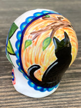 Load image into Gallery viewer, Inspired by nature, ceramic skull is full of vibrant colors and intricate details. The primary color is white with a black cat silhouette and multi-color floral motifs, along with hearts and leaves and lively colorful accents around the entire skull.  Mini skull represented the eternal love for a special kitty cat in your life. Black cat silhouette is sitting on a tree branch admiring the starry night sky. Art created for all the cat lovers who adore their sweet kitty.  Measurements: H 2 3/4” x W 2” x D 2”
