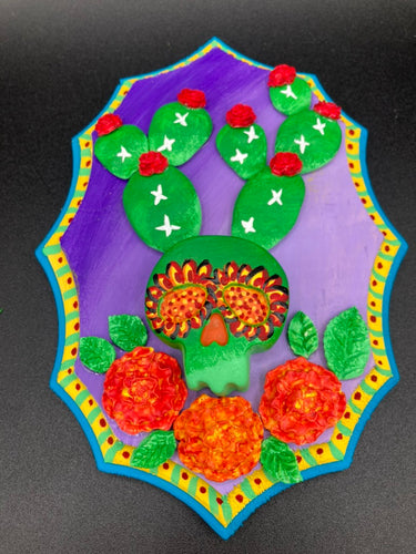 This handcrafted polymer clay wall art features a wood frame painted in a gradient from light to dark purple, creating a stunning sunset look. With a bright green skull cactus plant and bright red blooming flowers, this piece is sure to make a statement. Measurements: H 7