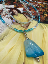 Load image into Gallery viewer, This exquisite necklace features a vibrant Sea Blue Agate Natural Stone Pendant and two strands of multi-colored glass beaded necklace. An adjustable collar-style strand contains small, light blue beads and lots of iridescent double-hole beads for shimmer and sparkle. The longer strand features larger blue glass beads interspersed with clear and silver beads for a sophisticated, draped look. It's finished with a set of dainty matching earrings for a complete look. Necklace is 14”adjustable up to 18"

