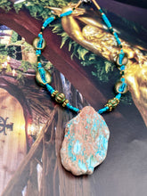 Load image into Gallery viewer, This stunning two-strand necklace features an array of glass beads in hues of green and blue turquoise, gold and green for a vibrant, colorful and bold look. The centerpiece is a large howlite stone chip dyed in turquoise color, accented by the bronze patina cowry beads.  Measurements: Necklace is 17” 
