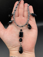 Load image into Gallery viewer, This adjustable collar-style necklace features silver and black glass beads in a combination of both teardrop and rectangular shapes, designed to create visual complexity and depth. The two strands are held together with two-hole silver and black beads, creating a stunningly bold and elegant draped collar that enhances any outfit.

