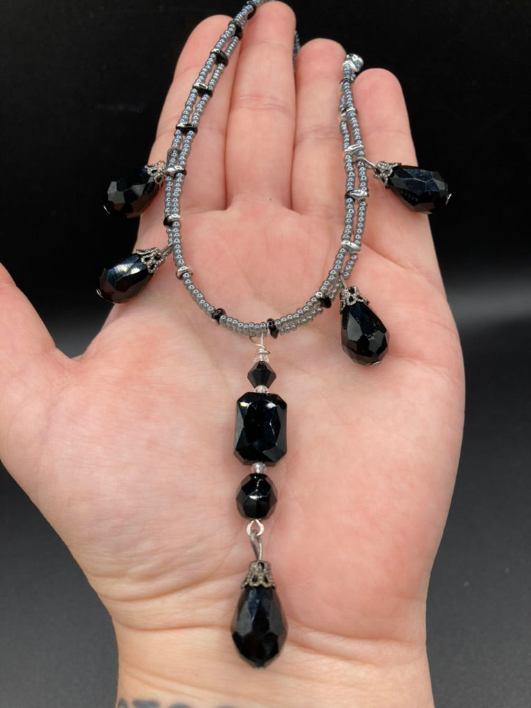 This adjustable collar-style necklace features silver and black glass beads in a combination of both teardrop and rectangular shapes, designed to create visual complexity and depth. The two strands are held together with two-hole silver and black beads, creating a stunningly bold and elegant draped collar that enhances any outfit.