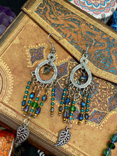 Load image into Gallery viewer, These large chandelier-style earrings feature blues, greens, and iridescent glass beads as well as a finely crafted silver plated leaf charm for added detail. Perfect for bohemian-inspired looks, these versatile earrings can elevate any wardrobe from whimsical hippie to boho chic.  Measurements: 3 1/4” in length
