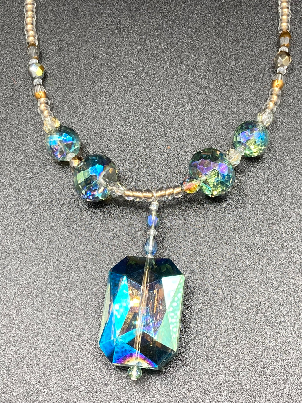 Make a bold statement with this shimmery aqua blue and gold tones glass beaded necklace. The large rectangular faceted shaped pendant creates a vivid contrast to the warm gold tones of the necklace, perfect for adding a rich pop of color to any Spring outfit. The draped design gives this piece an elegant, bohemian look. Measurements:    Necklace is 20” in length, 