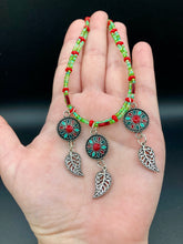 Load image into Gallery viewer, This adjustable necklace features an eye-catching Southwest-inspired design. Multi-hued glass beads, including bright green and vibrant red, are accompanied by dark red and green pendants with silver-plated feather charms. Ideal for chingona guerrera carbonas, la India princesses, warrior cosplay, and native-inspired looks.  Measurements:  Necklace is 15” adjustable up to 17" in length
