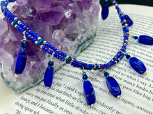 Load image into Gallery viewer, This Vibrant Royal Blue Glass Beaded Choker Necklace features two adjustable strands of deep blue beads with purple hues. The Bohemian style jewelry includes a mixture of rectangular-shaped blue, purple, and silver glass beads, which come together to create an eye-catching and elegant look. Perfect for any outfit, this draping necklace will add a vibrant and bold touch to your style.  Measurements:    Necklace is 14 3/4” in length, can be adjusted up to 16"
