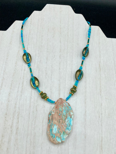 This stunning two-strand necklace features an array of glass beads in hues of green and blue turquoise, gold and green for a vibrant, colorful and bold look. The centerpiece is a large howlite stone chip dyed in turquoise color, accented by the bronze patina cowry beads.  Measurements: Necklace is 17” 