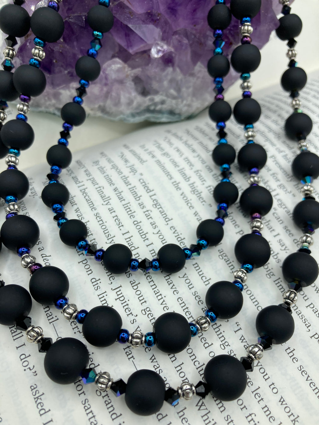 This statement piece features 3 long strands of flat matte black and dark blue glass beads, complemented by a floral metal beaded necklace. It gracefully drapes across the neck for a fierce, yet elegant look, making it perfect for cocktail chic attire, weddings, or minimalist styling.  Measurements:    Necklace is 16” in length - adjustable to 18