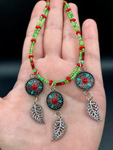 Load image into Gallery viewer, This adjustable necklace features an eye-catching Southwest-inspired design. Multi-hued glass beads, including bright green and vibrant red, are accompanied by dark red and green pendants with silver-plated feather charms. Ideal for chingona guerrera carbonas, la India princesses, warrior cosplay, and native-inspired looks.  Measurements:  Necklace is 15” adjustable up to 17" in length
