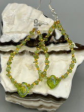 Load image into Gallery viewer, These natural peridot chips earrings feature a gold glass beading on their hoops, creating an eye-catching light green tone. A pressed glass leaf charm is also included, boasting an iridescent shimmer finish with subtle hints of gold, purple, blue, and pink. Measurements: 2 1/2&quot; length
