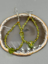 Load image into Gallery viewer, These natural peridot chips earrings feature a gold glass beading on their hoops, creating an eye-catching light green tone. A pressed glass leaf charm is also included, boasting an iridescent shimmer finish with subtle hints of gold, purple, blue, and pink.  Measurements: 2 1/2&quot; length
