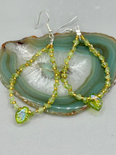 Load image into Gallery viewer, These natural peridot chips earrings feature a gold glass beading on their hoops, creating an eye-catching light green tone. A pressed glass leaf charm is also included, boasting an iridescent shimmer finish with subtle hints of gold, purple, blue, and pink. Measurements: 2 1/2&quot; length
