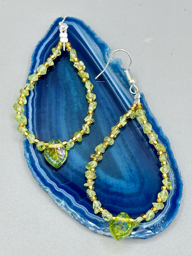 These natural peridot chips earrings feature a gold glass beading on their hoops, creating an eye-catching light green tone. A pressed glass leaf charm is also included, boasting an iridescent shimmer finish with subtle hints of gold, purple, blue, and pink. Measurements: 2 1/2