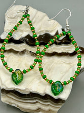 Load image into Gallery viewer, This pair of earrings features a dark green, pressed glass leaf charm in an iridescent finish that gives off subtle hues of gold, purple, blue, and pink. Complemented with green and gold glass beaded hoop earrings, this set is sure to sparkle. Measurements: 2 3/4&quot; length
