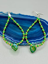 Load image into Gallery viewer, This pair of earrings features a dark green, pressed glass leaf charm in an iridescent finish that gives off subtle hues of gold, purple, blue, and pink. Complemented with green and gold glass beaded hoop earrings, this set is sure to sparkle. Measurements: 2 3/4&quot; length
