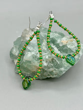 Load image into Gallery viewer, This pair of earrings features a dark green, pressed glass leaf charm in an iridescent finish that gives off subtle hues of gold, purple, blue, and pink. Complemented with green and gold glass beaded hoop earrings, this set is sure to sparkle. Measurements: 2 3/4&quot; length
