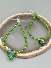 Load image into Gallery viewer, This pair of earrings features a dark green, pressed glass leaf charm in an iridescent finish that gives off subtle hues of gold, purple, blue, and pink. Complemented with green and gold glass beaded hoop earrings, this set is sure to sparkle. Measurements: 2 3/4&quot; length
