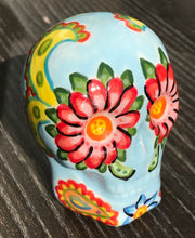 Load image into Gallery viewer, This ceramic skull statuette is a stunning piece of art inspired by nature. Its soft periwinkle blue hue is decorated with multi-color floral motifs, hearts, leaves and colorful accents, creating a beautiful piece of hand-painted, Day of the Dead-themed art.  H 2 3/4” x W 2” x D 2”
