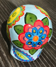 Load image into Gallery viewer, Inspired by nature, this ceramic skull statuette is full of vibrant color and intricate details. The primary color is soft, light periwinkle blue with Multi-color floral motifs that are painted across the skull, along with hearts and leaves and lively colorful accents around the entire skull.
