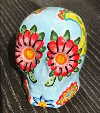 Load image into Gallery viewer,  Inspired by nature, this ceramic skull statuette is full of vibrant color and intricate details. The primary color is soft, light periwinkle blue with Multi-color floral motifs that are painted across the skull, along with hearts and leaves and lively colorful accents around the entire skull.

