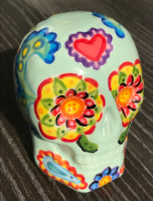 Load image into Gallery viewer, Inspired by nature, this ceramic skull statuette is full of vibrant color and intricate details. The primary color is soft pastel green with Multi-color floral motifs that are painted across the skull, along with hearts and leaves and lively colorful accents around the entire skull.
