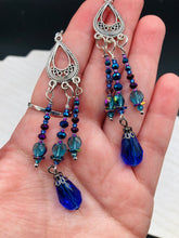 Load image into Gallery viewer, This set of large, eye-catching chandelier-style earrings draw inspiration from the vibrant array of colors found in the galaxy and cosmos. Featuring three strands of shimmering glass beads in bold shades of blue, purple, gold, and green, it's an essential piece for any jewelry collection.  Measurements: 3 1/2” in length
