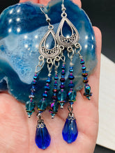 Load image into Gallery viewer, This set of large, eye-catching chandelier-style earrings draw inspiration from the vibrant array of colors found in the galaxy and cosmos. Featuring three strands of shimmering glass beads in bold shades of blue, purple, gold, and green, it's an essential piece for any jewelry collection.  Measurements: 3 1/2” in length
