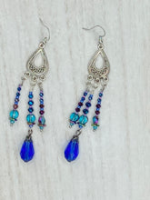 Load image into Gallery viewer, This set of large, eye-catching chandelier-style earrings draw inspiration from the vibrant array of colors found in the galaxy and cosmos. Featuring three strands of shimmering glass beads in bold shades of blue, purple, gold, and green, it's an essential piece for any jewelry collection.  Measurements: 3 1/2” in length

