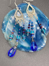 Load image into Gallery viewer, This set of large, eye-catching chandelier-style earrings draw inspiration from the vibrant array of colors found in the galaxy and cosmos. Featuring three strands of shimmering glass beads in bold shades of blue, purple, gold, and green, it's an essential piece for any jewelry collection.  Measurements: 3 1/2” in length
