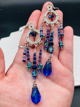Load image into Gallery viewer, This beautifully large and bold set of chandelier earrings were inspired by the vibrant and beautiful galaxy and cosmos full of bright and stunning hues of blues and purples.  I created this set of earrings with strands of glass beads full of shimmer and bold hues of blues, purples and a tint of gold and greens colors. This is a long, large, and bold piece of jewelry have a lot of shimmer and movement, a “must have” to your collection.   Measurements: 4” in length
