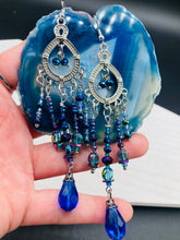 Load image into Gallery viewer, This beautifully large and bold set of chandelier earrings were inspired by the vibrant and beautiful galaxy and cosmos full of bright and stunning hues of blues and purples.  I created this set of earrings with strands of glass beads full of shimmer and bold hues of blues, purples and a tint of gold and greens colors. This is a long, large, and bold piece of jewelry have a lot of shimmer and movement, a “must have” to your collection.   Measurements: 4” in length

