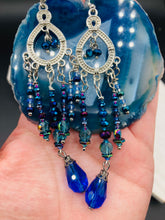 Load image into Gallery viewer, This beautifully large and bold set of chandelier earrings were inspired by the vibrant and beautiful galaxy and cosmos full of bright and stunning hues of blues and purples.  I created this set of earrings with strands of glass beads full of shimmer and bold hues of blues, purples and a tint of gold and greens colors. This is a long, large, and bold piece of jewelry have a lot of shimmer and movement, a “must have” to your collection.   Measurements: 4” in length
