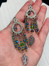 Load image into Gallery viewer, These large chandelier-style earrings feature blues, greens, and iridescent glass beads as well as a finely crafted silver plated leaf charm for added detail. Perfect for bohemian-inspired looks, these versatile earrings can elevate any wardrobe from whimsical hippie to boho chic.  Measurements: 3 1/4” in length
