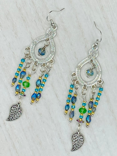 These large chandelier-style earrings feature blues, greens, and iridescent glass beads as well as a finely crafted silver plated leaf charm for added detail. Perfect for bohemian-inspired looks, these versatile earrings can elevate any wardrobe from whimsical hippie to boho chic.  Measurements: 3 1/4” in length
