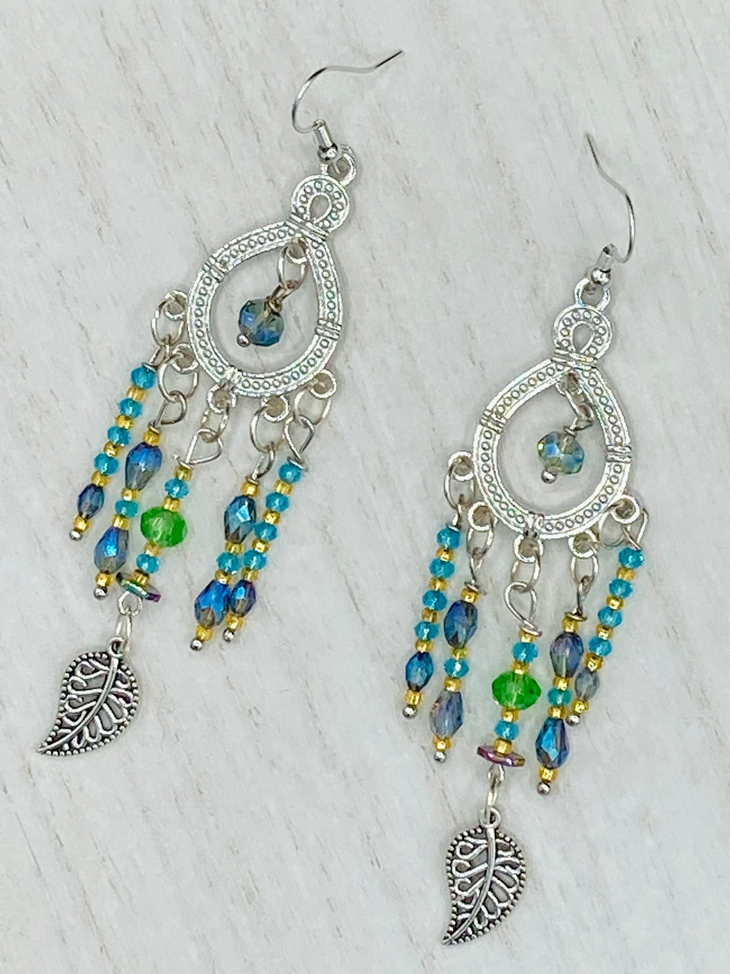 These large chandelier-style earrings feature blues, greens, and iridescent glass beads as well as a finely crafted silver plated leaf charm for added detail. Perfect for bohemian-inspired looks, these versatile earrings can elevate any wardrobe from whimsical hippie to boho chic.  Measurements: 3 1/4” in length
