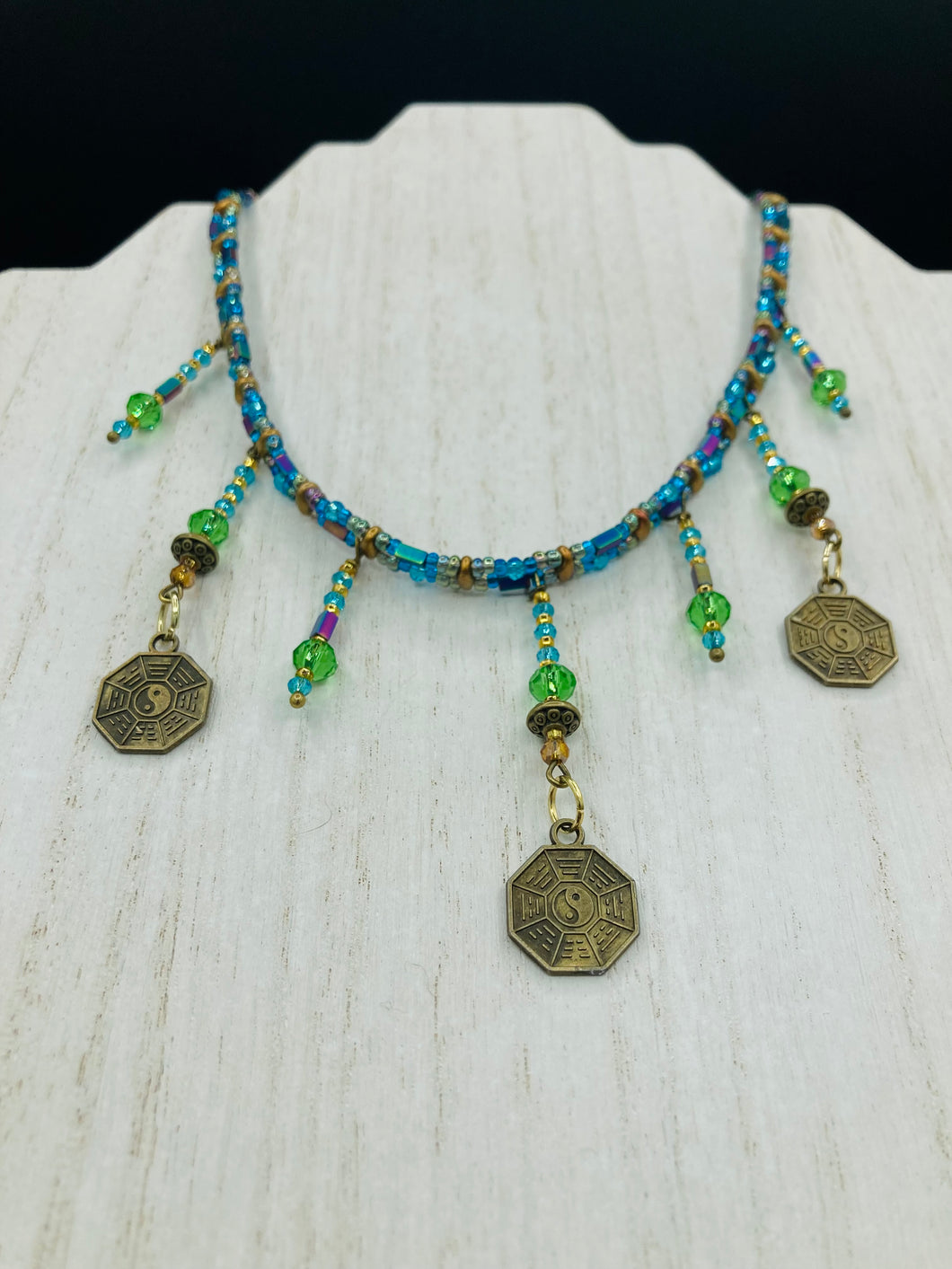This collar-style necklace is a showstopper, beaded with stylish glass beads in green and blue tones, featuring contrasting yin-yang charms with differing sides. Rectangular rainbow hematite beads add complexity and depth, and double-hole matte gold beads keep the collar shape complete. It gracefully drapes around the neck, making a timeless statement of elegance.  Measurements:  Necklace is 14”, can be adjusted up to 18