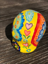 Load image into Gallery viewer, Inspired by nature, this ceramic skull statuette is full of vibrant color and intricate details. The primary color is yellow with Multi-color floral motifs that are painted across the skull, along with hearts and leaves and lively colorful accents around the entire skull.
