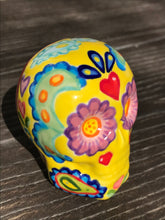 Load image into Gallery viewer, Inspired by nature, this ceramic skull statuette is full of vibrant color and intricate details. The primary color is yellow with Multi-color floral motifs that are painted across the skull, along with hearts and leaves and lively colorful accents around the entire skull.
