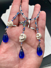 Load image into Gallery viewer, This adjustable collar-style necklace features two shimmery strands of silver and blue glass beads, with three skull charms boasting large, luminous blue teardrop glass beads adding extra shimmer and movement.  Measurements:  Necklace is 14 3/8” adjustable up to 18" in length
