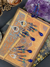 Load image into Gallery viewer, This set of large, eye-catching chandelier-style earrings draw inspiration from the vibrant array of colors found in the galaxy and cosmos. Featuring three strands of shimmering glass beads in bold shades of blue, purple, gold, and green, it's an essential piece for any jewelry collection.  Measurements: 3 1/2” in length
