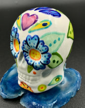 Load image into Gallery viewer, Inspired by nature, this ceramic skull statuette is full of vibrant color and intricate details that I hand painted. The primary color is white with multi-color floral motifs that are painted across the skull, along with hearts and leaves and lively colorful accents around the entire skull. 

