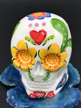 Load image into Gallery viewer, Inspired by nature, this ceramic skull statuette is full of vibrant color and intricate details. The primary color is white with multi-color floral motifs that are painted across the skull, along with hearts and leaves and lively colorful accents around the entire skull.
